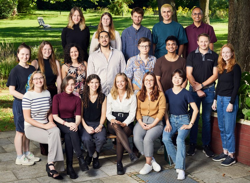 Unsere fünfte Kohorte (Start 2023) Eine Gruppe von Menschen, die in Freizeitkleidung in einem Park posiert. Im Hintergrund sind Liegestühle und Bäume zu sehen, die eine entspannte Atmosphäre schaffen. Die Personen sitzen und stehen in zwei Reihen.