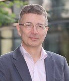 Person in Geschäftskleidung vor einem Bürogebäude, mit lockigem Haar, grauem Anzug und rosa Hemd, im Freien.