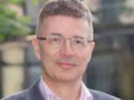 Person in Geschäftskleidung vor einem Bürogebäude, mit lockigem Haar, grauem Anzug und rosa Hemd, im Freien.