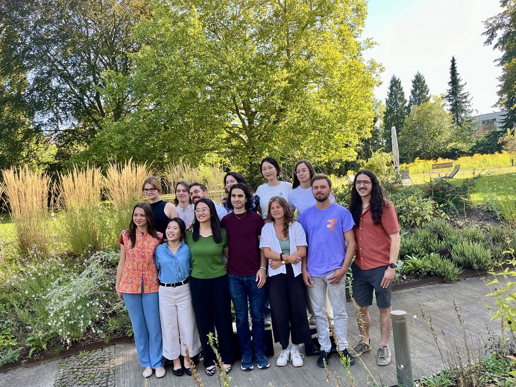 A diverse group of individuals stands closely together in a garden, with tall grass and trees visible in the background, suggesting a warm, sunny day.