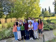 A diverse group of individuals stands closely together in a garden, with tall grass and trees visible in the background, suggesting a warm, sunny day.