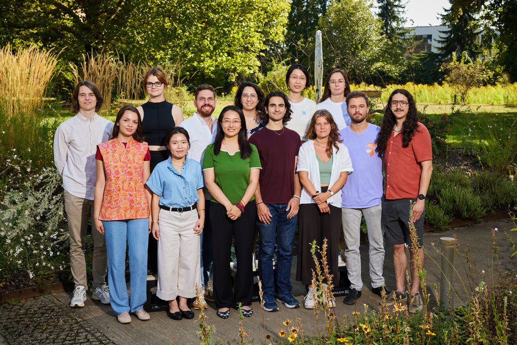 A mixed-gender group poses outdoors in casual clothing, standing on pavement beside a grassy area, with trees and lounge chairs in the background.