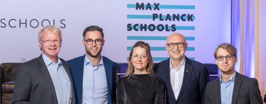 Unsere Mission - die Max Planck Schools Fünf Personen stehen nebeneinander, formell gekleidet, vor einem Banner der Max Planck Schools, das von türkisen Streifen akzentuiert wird.