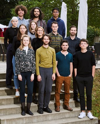 Carolin, Leonardo, Rebekka, Bojana, Nina, Hassan, Karla, Pietro, Oliver, Pat, Pietro, Caedyn und Moritz (left to right) A diverse group of people stands arranged on exterior steps. They are dressed in casual attire, with trees and patio chairs in the background.
