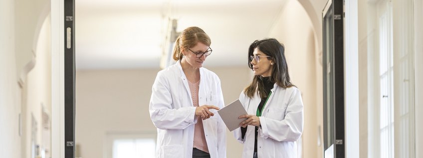 Clinician Scientist Program. - Next Application Period from September 1st to December 1st 2026 Two researchers in white lab coats converse while examining a document in a brightly lit hallway.