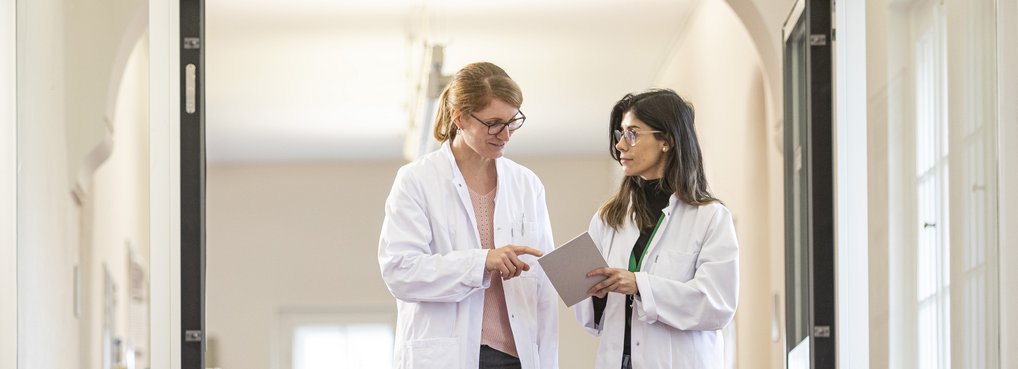 Zwei Personen in weißen Laborkitteln stehen in einem hellen Flur und betrachten ein Tablet.