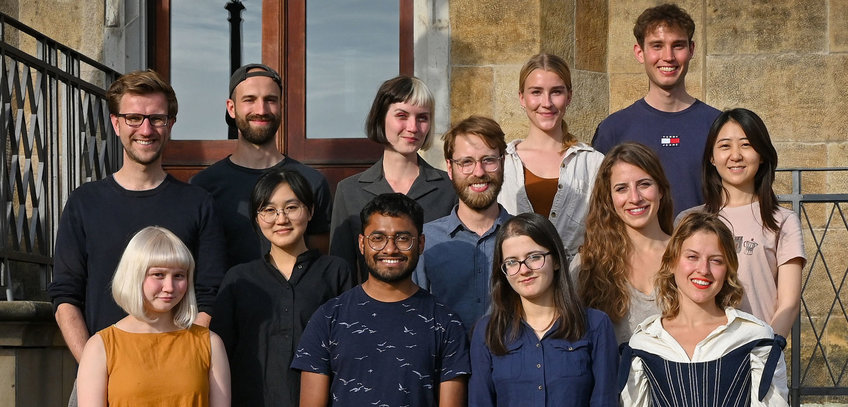 Unsere dritte Kohorte (Start 2021) Zwölf Menschen in verschiedenen Freizeitkleidungen stehen auf einer Treppe vor einem historischen Gebäude mit Holztür.