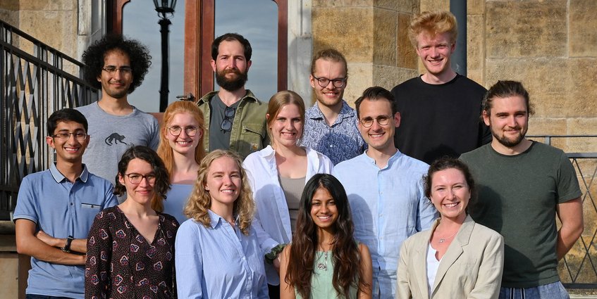 Unsere zweite Kohorte (Start 2020) Eine Gruppe von Personen, bestehend aus Männern und Frauen, posiert vor einem historischen Gebäude mit markanten Sandsteinmauern und einem dekorativen Eisengeländer. Im Hintergrund ist eine Straßenlaterne zu sehen.