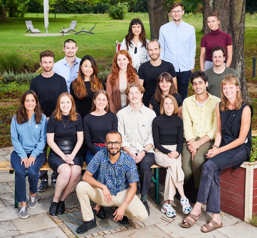 Unsere vierte Kohorte (Start 2022) Eine Gruppe von Menschen, die in Freizeitkleidung in einem Park posiert. Im Hintergrund sind Liegestühle und Bäume zu sehen, die eine entspannte Atmosphäre schaffen. Die Personen sitzen und stehen in zwei Reihen.